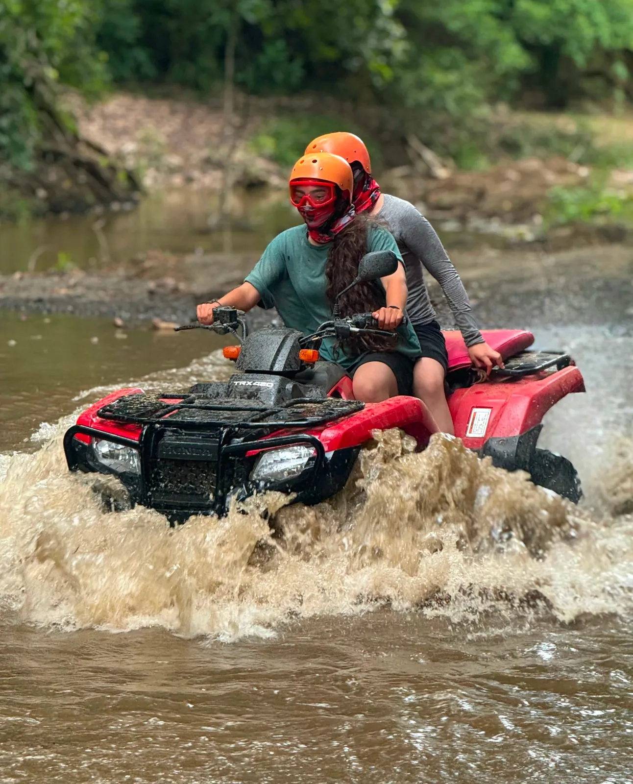 Two riders in helmets and goggles splashing through a river crossing on a red Honda ATV in Guanacaste