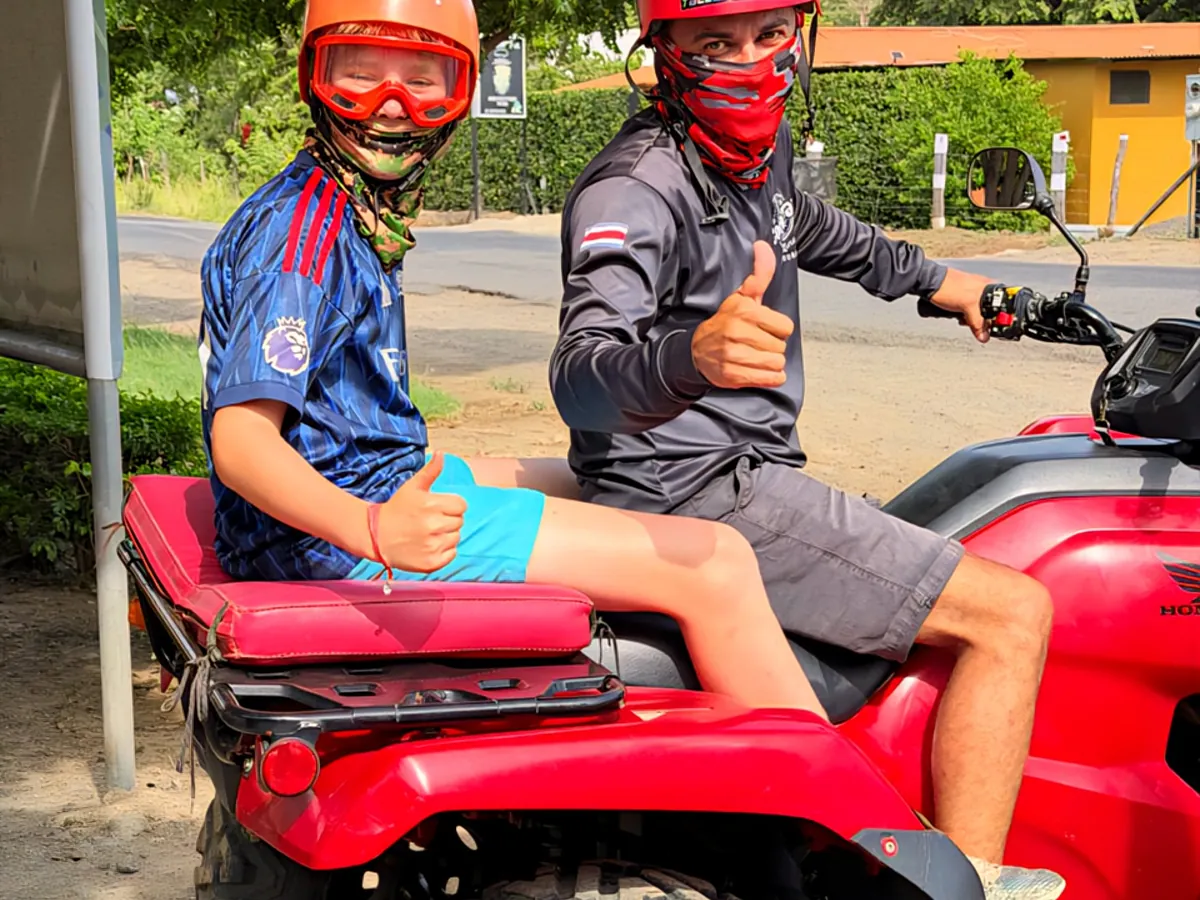 A young passenger riding tandem behind a guide on a red Honda ATV, both giving a thumbs up