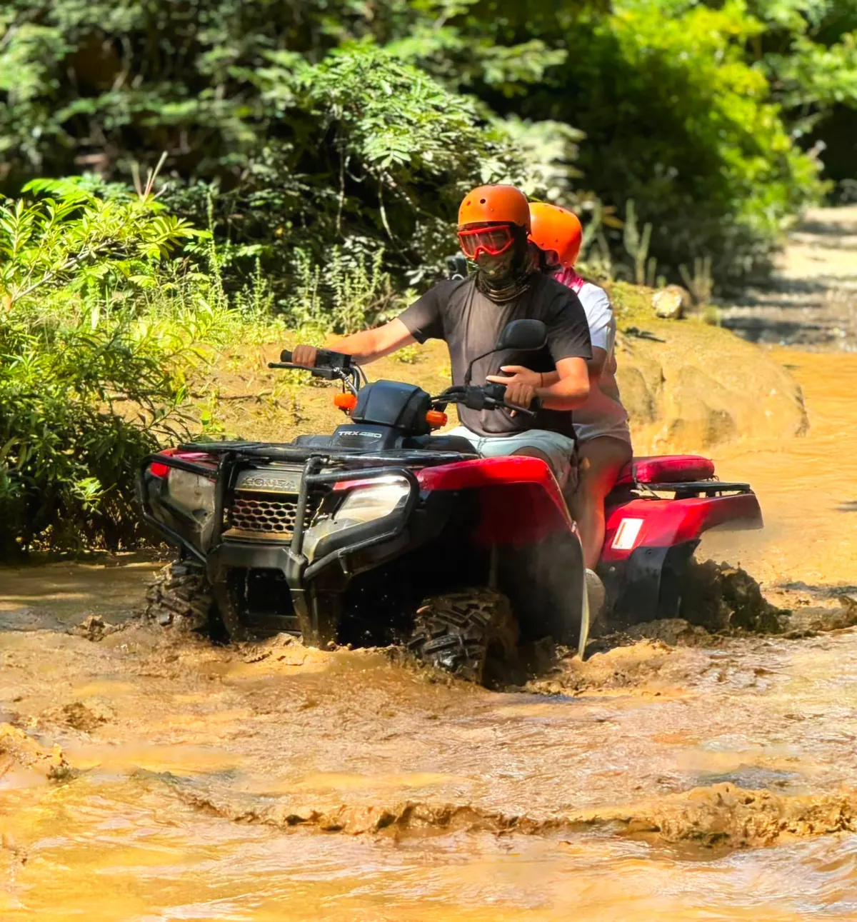 Two riders on a red Honda ATV splashing through a muddy river crossing on the way to a waterfall