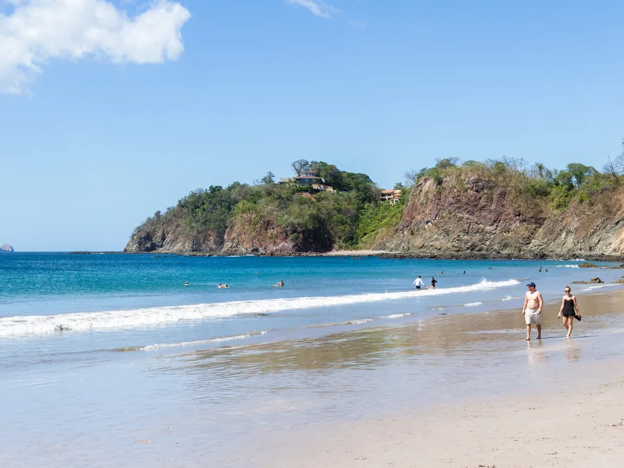Couple walking a sandy Pacific beach below a rocky, forested headland in Guanacaste