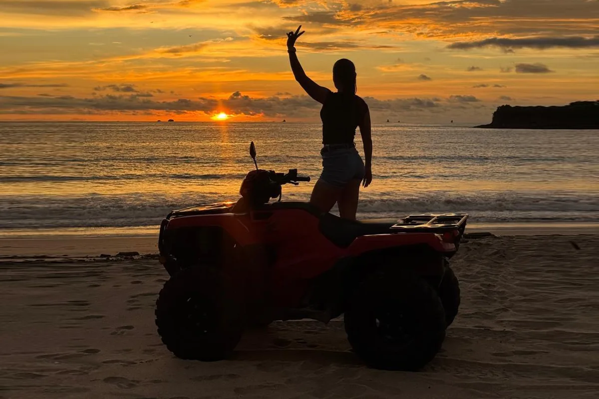 Rider standing on a red ATV on the beach at sunset near Playas del Coco, waving toward the Pacific