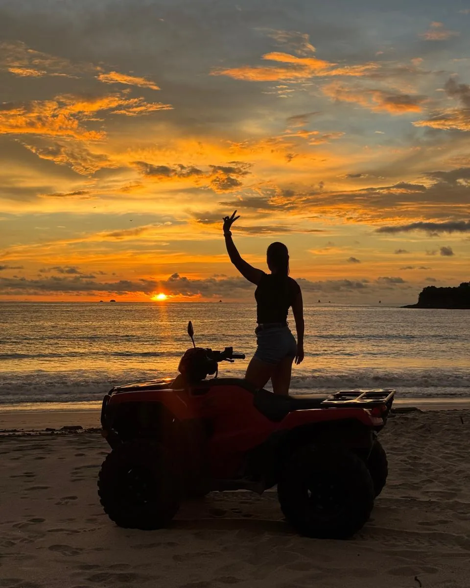 Rider standing on a red ATV on the beach at sunset near Playas del Coco, waving toward the Pacific